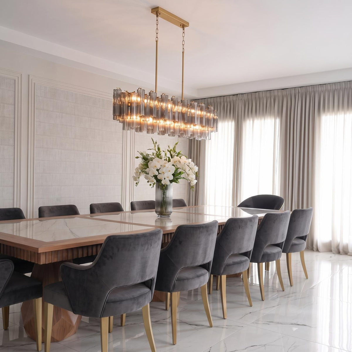 A contemporary dining room with a brushed brass linear chandelier featuring ribbed smoke glass rods over a marble table with gray velvet chairs and paneled walls.