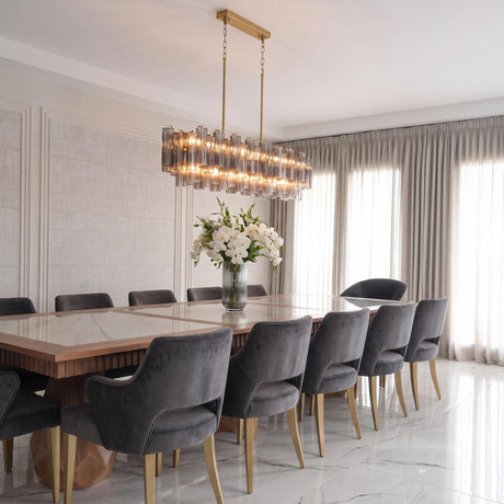 A contemporary dining room with a brushed brass linear chandelier featuring ribbed smoke glass rods over a marble table with gray velvet chairs and paneled walls.