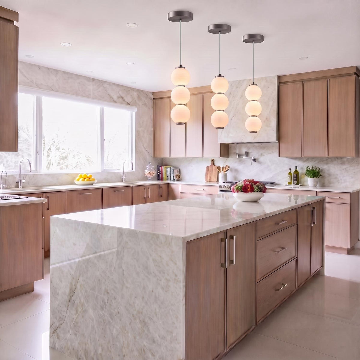 A contemporary kitchen with three brushed nickel and opal glass globe pendants suspended over a large white marble waterfall island surrounded by light wood cabinetry.