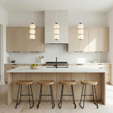A minimalist kitchen featuring three Shai LED Pendant 7 inch Matte Black and Opal Glass pendants over a white island with light oak cabinetry and four wooden stools.