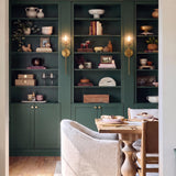 A transitional dining room featuring the Gurt 1 Light Wall Sconce 20", Aged Brass mounted on deep green shelving behind a light wood table with upholstered chairs.