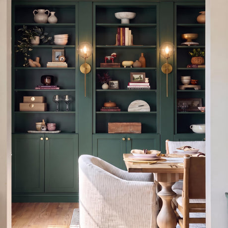 A transitional dining room featuring the Gurt 1 Light Wall Sconce 20", Aged Brass mounted on deep green shelving behind a light wood table with upholstered chairs.