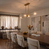 A transitional dining room with the Gurt 4 Light Linear Pendant 48 in aged brass featuring glass globes over a walnut dining table and cream chairs.