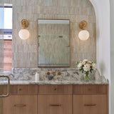 A transitional bathroom featuring two Tristen Wall Sconce 12 inch fixtures in brushed brass and opal glass flanking a mirror over a marble countertop and fluted cabinets.
