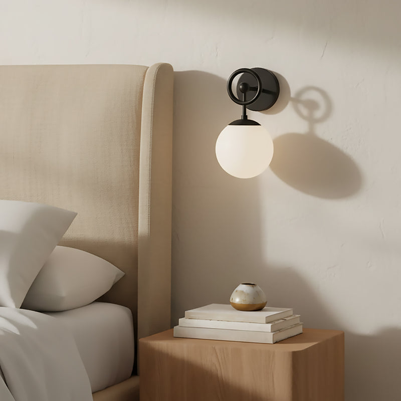 Tristen wall-mounted lamp in black and opal glass, casting a soft glow with a bed and nightstand in the foreground.