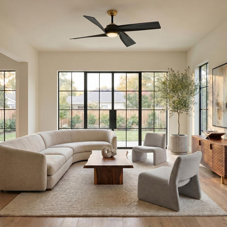 A contemporary living room with a brushed brass and matte black ceiling fan centered above a curved beige sectional and large black-framed glass doors.