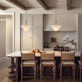 A modern farmhouse kitchen with two brushed brass pendants featuring frosted glass cone shades suspended over a white marble island and woven bar stools beneath a beamed ceiling.