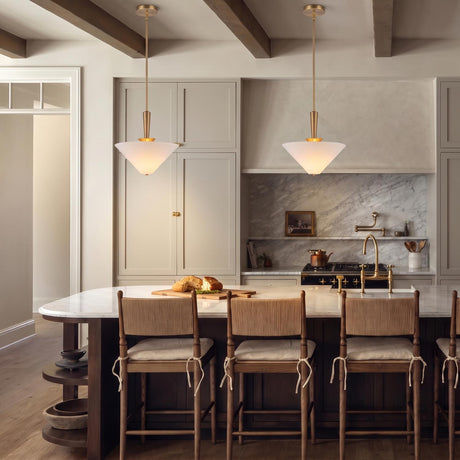 A modern farmhouse kitchen with two brushed brass pendants featuring frosted glass cone shades suspended over a white marble island and woven bar stools beneath a beamed ceiling.