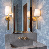 A transitional bathroom with two Sabina Wall Sconce 18-inch brushed brass fixtures featuring white linen shades flanking a gold framed mirror above a gray marble sink.