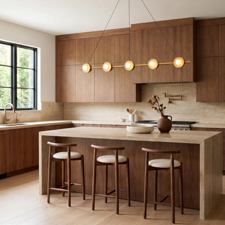 A modern kitchen with the Sol LED Linear Pendant 48", Brushed Brass suspended over a beige stone waterfall island featuring walnut cabinets and three wood bar stools.