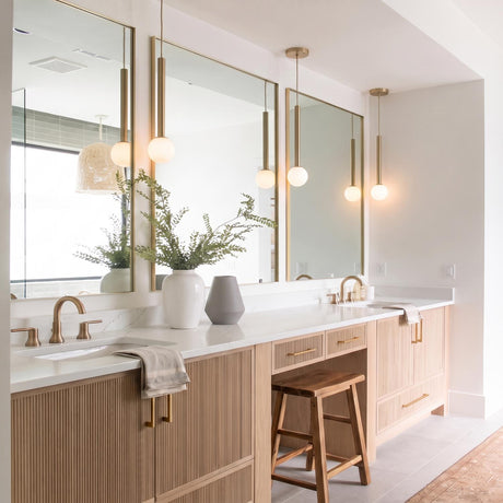 A modern bathroom featuring four brushed brass globe pendants with frosted glass shades hanging above a light wood slatted vanity and white quartz countertops.