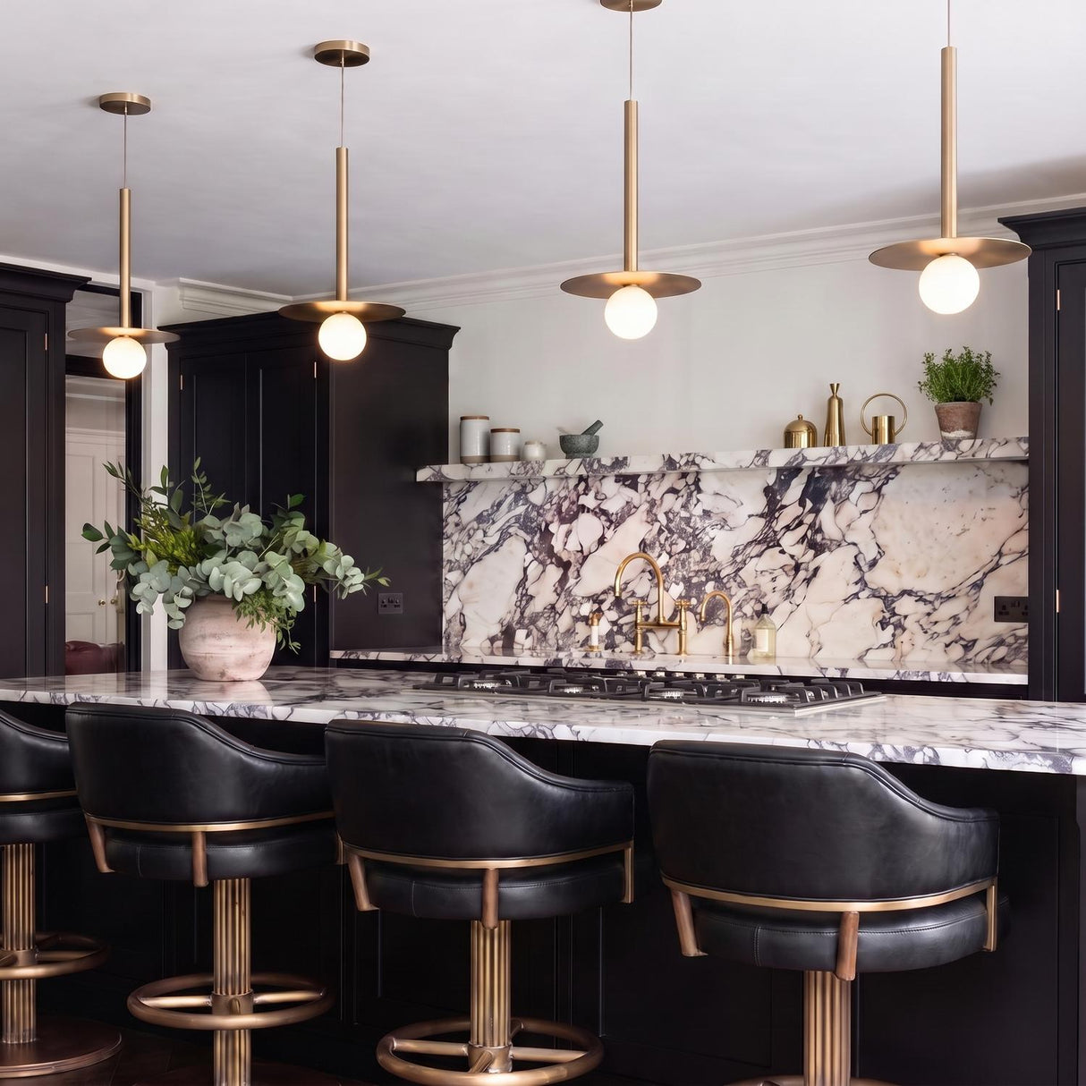A modern kitchen featuring four brushed brass saucer pendants over a purple-veined white marble island with black cabinetry and matching black leather bar stools.