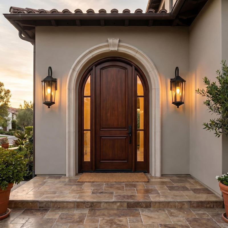A Mediterranean stucco entry featuring two matte black Asbury Outdoor Wall Lights flanking an arched mahogany door at dusk, with a stone tile porch and greenery.