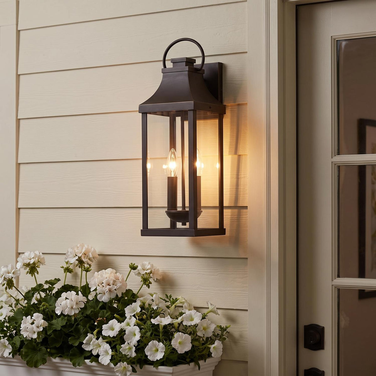 A cream-sided porch entry features the glowing Ainsley Outdoor Wall Light, Medium 21" Oiled Bronze lantern beside a glass door above white potted flowers at dusk.