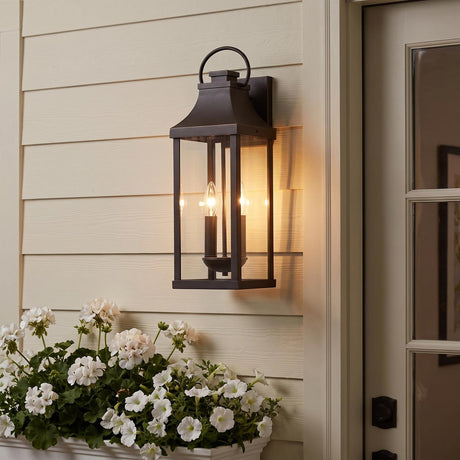 A cream-sided porch entry features the glowing Ainsley Outdoor Wall Light, Medium 21" Oiled Bronze lantern beside a glass door above white potted flowers at dusk.