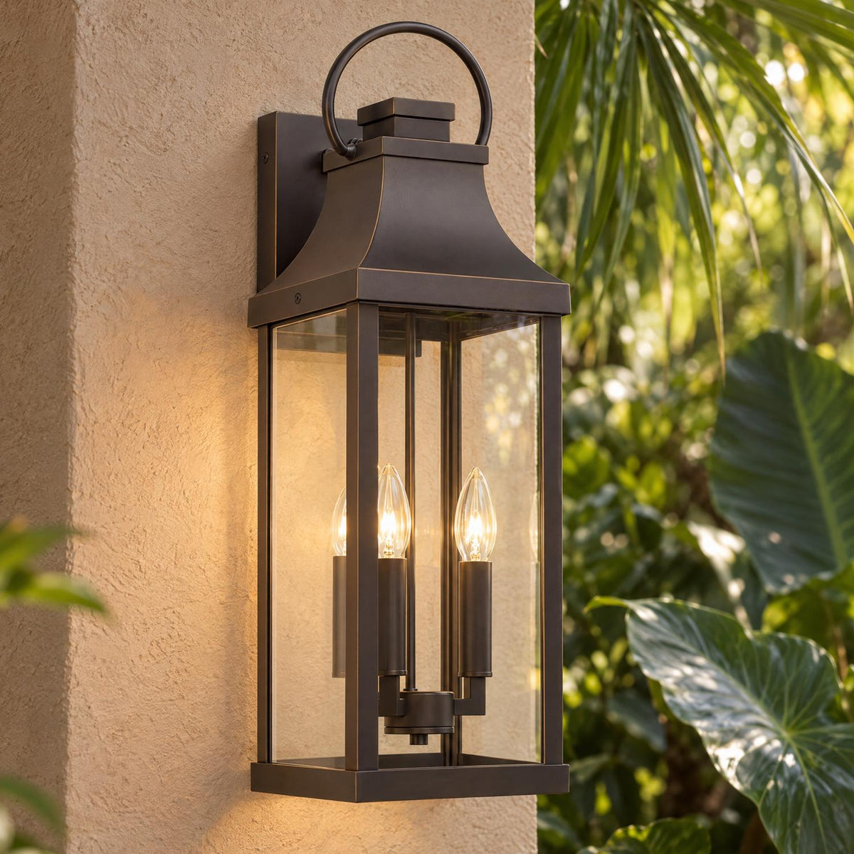 A Mediterranean home exterior features an Ainsley Outdoor Wall Light, Large 24" Oiled Bronze glowing on a beige stucco pillar framed by tropical foliage.