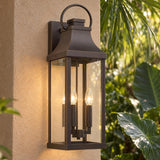 A Mediterranean home exterior features an Ainsley Outdoor Wall Light, Large 24" Oiled Bronze glowing on a beige stucco pillar framed by tropical foliage.
