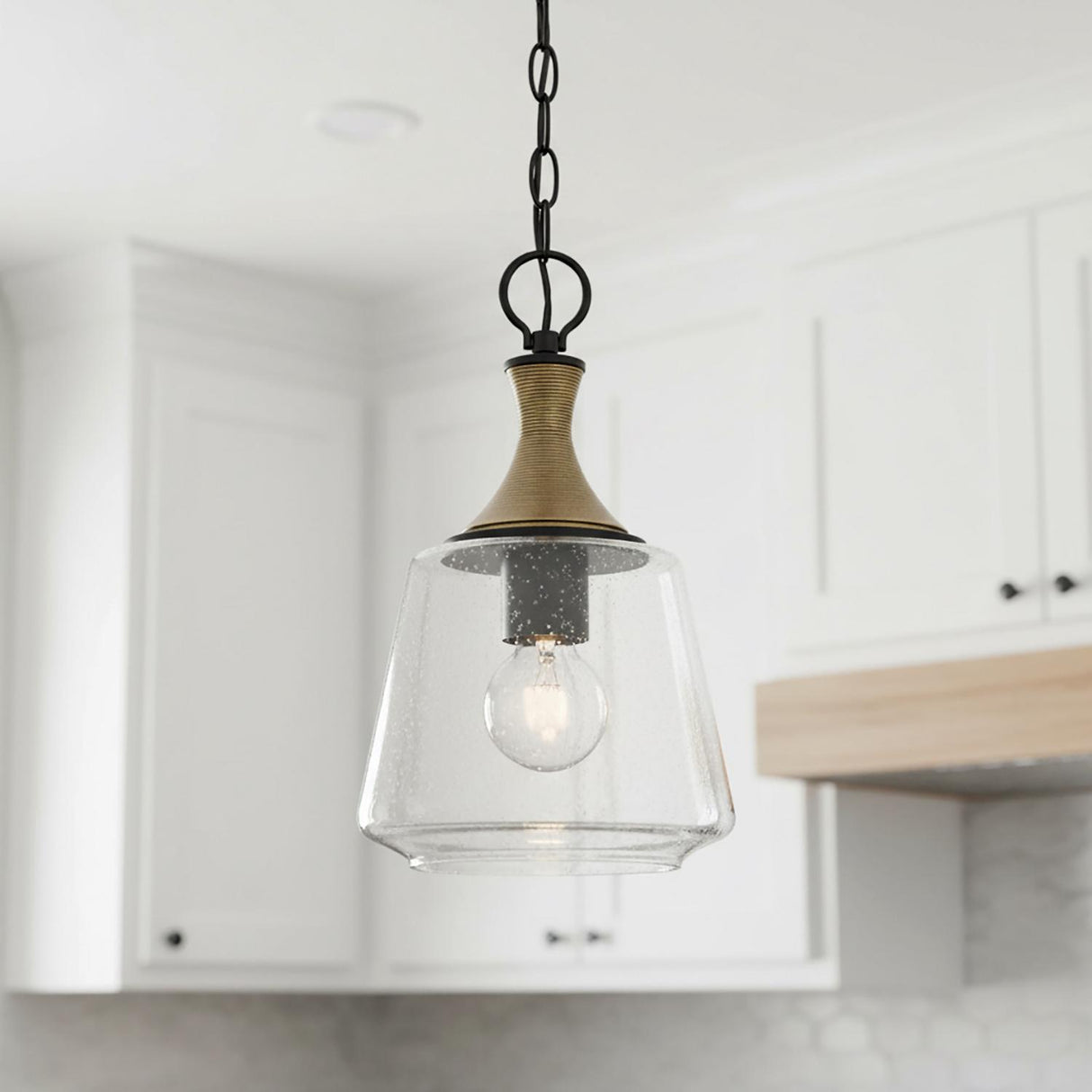 A transitional kitchen features the Malin Pendant 9", Matte Black with Brass Accents with its seeded glass shade suspended over a counter near white shaker cabinets.