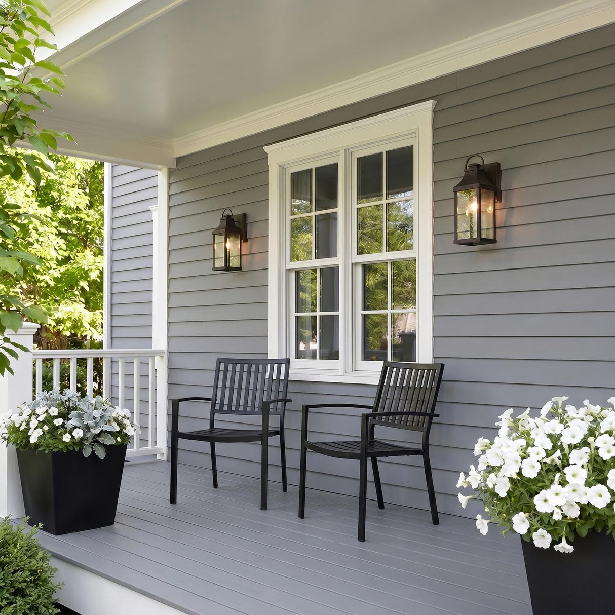 A modern farmhouse porch with two oiled bronze wall lanterns glowing at dusk, flanking a white window on gray siding with black chairs and white flowers.
