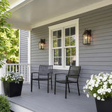 A modern farmhouse porch with two oiled bronze wall lanterns glowing at dusk, flanking a white window on gray siding with black chairs and white flowers.