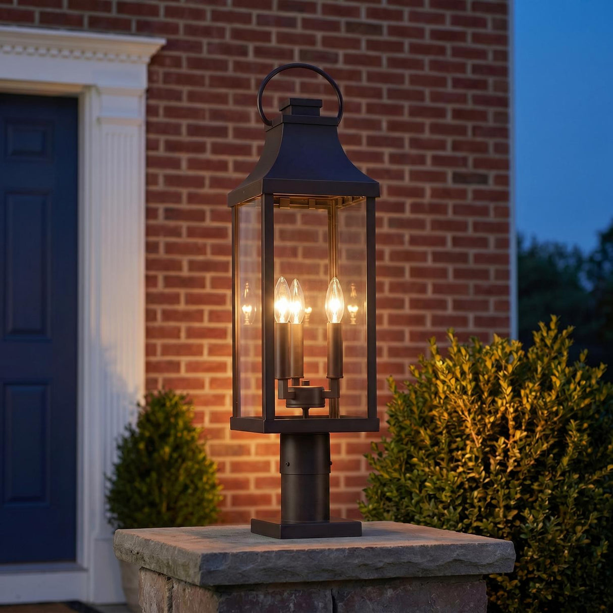 A traditional outdoor entryway featuring an Ainsley Outdoor Post Light, Oiled Bronze with clear glass panels mounted on a stone pier beside a red brick wall.
