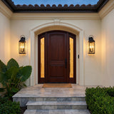 A Mediterranean entryway with two matte black Ainsley Outdoor Wall Lights flanking a dark wood door on beige stucco at dusk, featuring stone steps and boxwood.