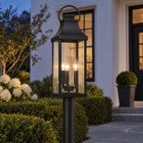 A modern farmhouse exterior features the glowing Ainsley Outdoor Post Light, Black along a stone pathway framed by manicured boxwood hedges and white brick at dusk.