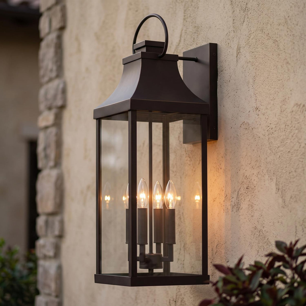 A Mediterranean home exterior featuring the Ainsley Outdoor Wall Light, Extra Large 27" Oiled Bronze lantern glowing at dusk on a stucco wall beside a stone corner.