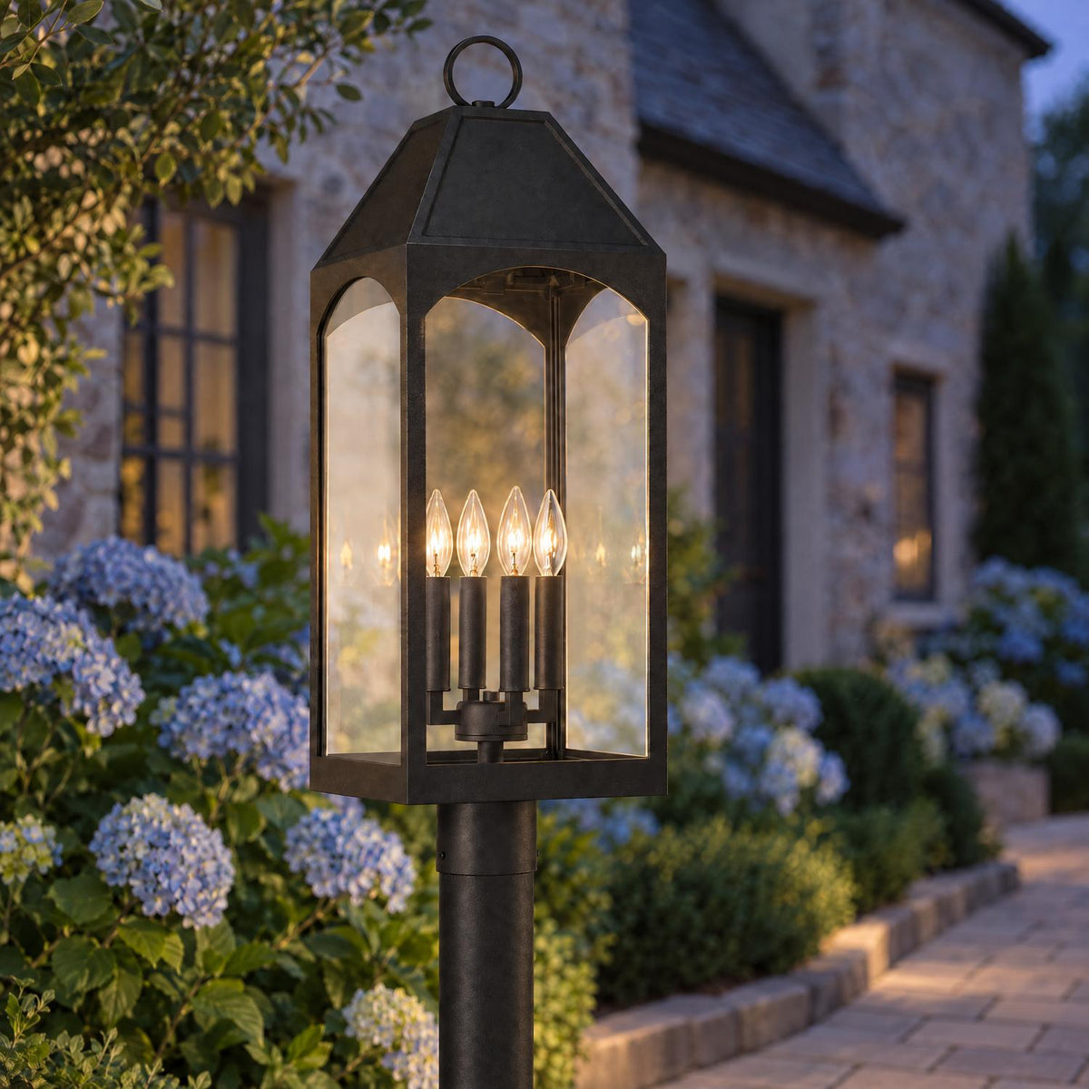 A traditional stone home exterior featuring the Chester Outdoor Post Light, Black glowing along a paver walkway surrounded by blue hydrangeas at dusk.