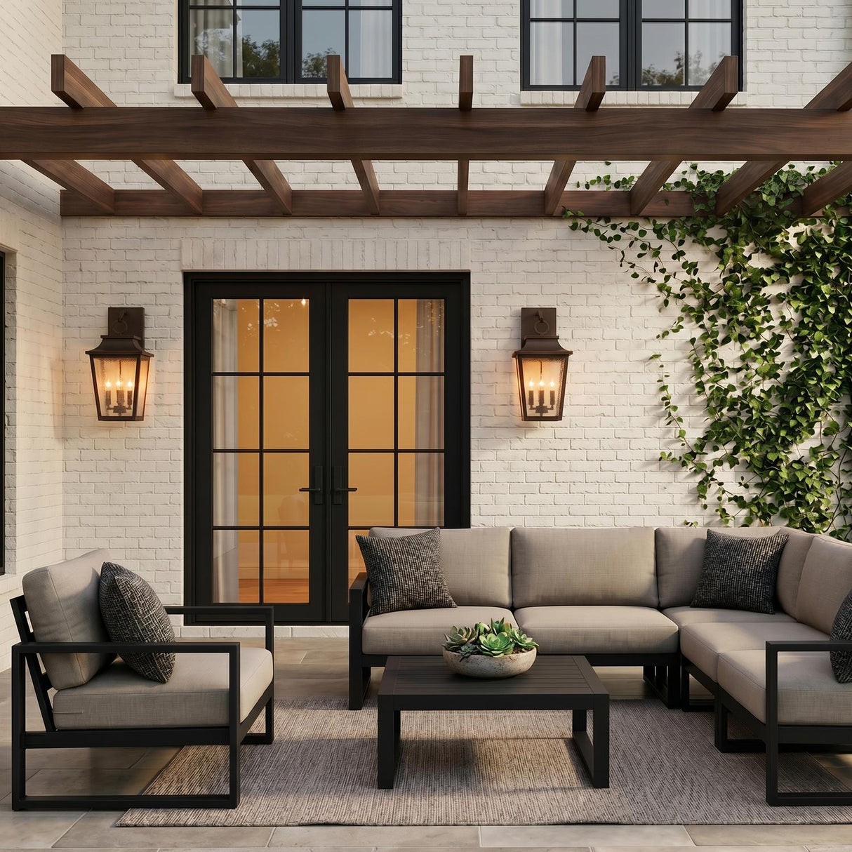 A modern farmhouse patio featuring the Beckett Outdoor Wall Light, Large 24" Oiled Bronze flanking black French doors on a white brick wall at dusk with climbing ivy.