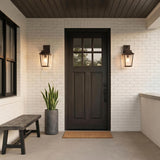 A modern farmhouse porch with two oiled bronze wall lanterns glowing at dusk, flanking a dark door on white brick beside a potted snake plant and bench.