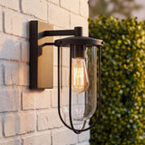 A modern farmhouse patio features the Orson Outdoor Wall Light, Small 12" Black, glowing in matte black against a white brick wall beside green hedges.