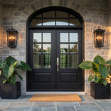 A traditional stone entry with two matte black wall lanterns flanking an arched black double door at dusk, featuring large potted elephant ear plants on slate tile.