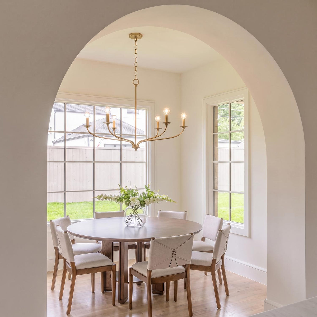 A transitional dining room with the Oriana 6 Light Chandelier 40", Matte Brass above a round walnut dining table featuring upholstered chairs and large grid windows.