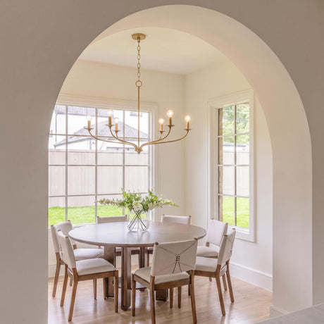 A transitional dining room with the Oriana 6 Light Chandelier 40", Matte Brass above a round walnut dining table featuring upholstered chairs and large grid windows.
