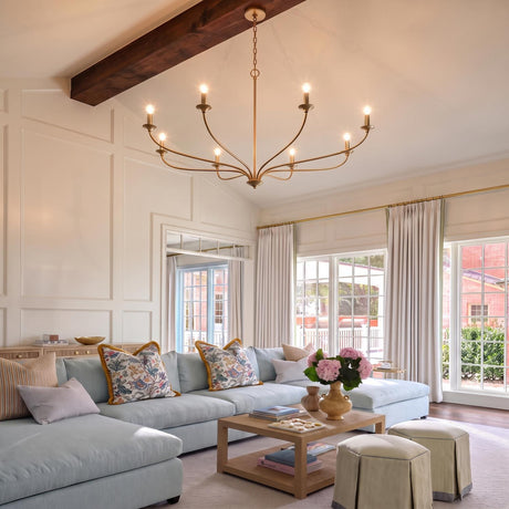A transitional living room with a matte brass Oriana 8 Light Chandelier hanging from a dark wood beam over a blue sectional sofa and white paneled walls.