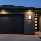 Orley Outdoor Wall Light, 16" tall in Black - A contemporary black wall light illuminates the exterior of a modern home next to a garage door.