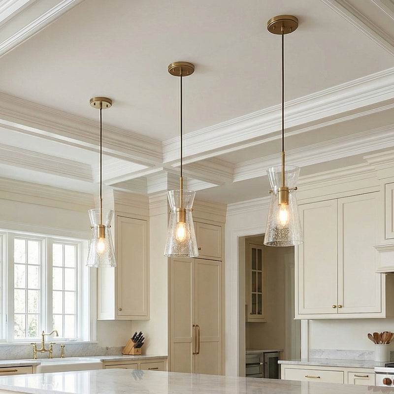 A traditional kitchen features three Aries Pendant 6", Aged Brass fixtures with seeded glass shades over the marble island under a white coffered ceiling and cream cabinets.