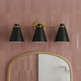 Avin 3 Light Vanity, Matte Black and Aged Brass - A three-light vanity fixture in matte black and aged brass hangs above an arched mirror, mounted on a tiled wall.