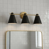 Avin 3 Light Vanity, Matte Black and Aged Brass - Three black conical shades with aged brass accents are mounted on a linear bar above a gold-framed mirror, all against a backdrop of white tiled walls.