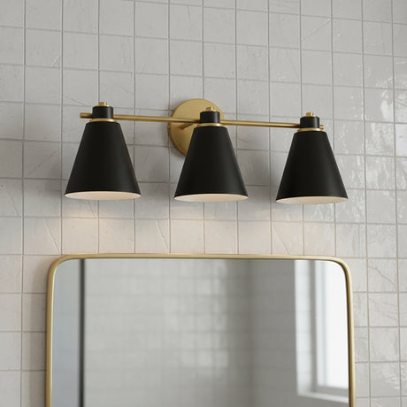 Avin 3 Light Vanity, Matte Black and Aged Brass - Three black conical shades with aged brass accents are mounted on a linear bar above a gold-framed mirror, all against a backdrop of white tiled walls.