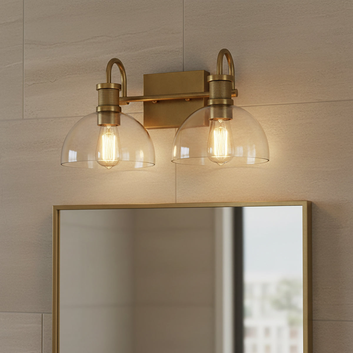Greer 2 Light Vanity, Aged Brass - Two glass dome shades with exposed Edison bulbs mounted on an aged brass fixture above a mirror with an aged brass frame, against a tiled wall.