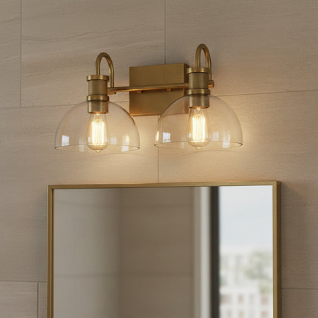 Greer 2 Light Vanity, Aged Brass - Two glass dome shades with exposed Edison bulbs mounted on an aged brass fixture above a mirror with an aged brass frame, against a tiled wall.