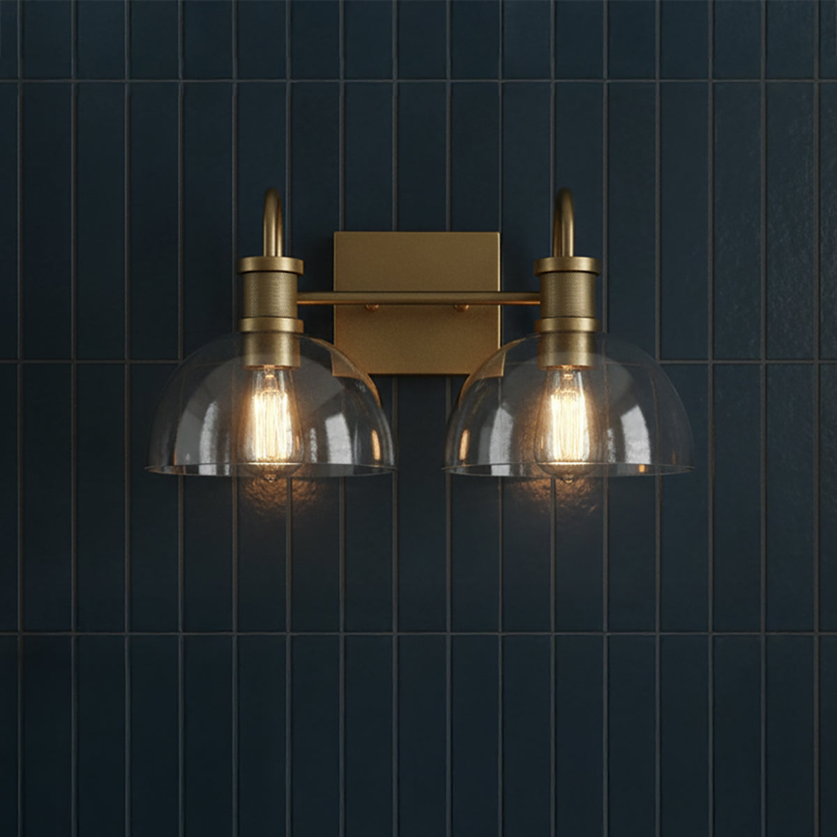 Greer 2 Light Vanity, Aged Brass - A two-light aged brass vanity fixture with clear glass dome shades hangs against a dark blue vertical tiled wall.