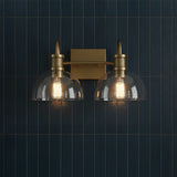 Greer 2 Light Vanity, Aged Brass - A two-light aged brass vanity fixture with clear glass dome shades hangs against a dark blue vertical tiled wall.