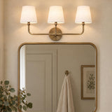 A transitional bathroom with the Elowen 3 Light Vanity, Aged Brass featuring white fabric shades mounted above a rounded brass mirror and eucalyptus.