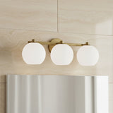 Enid 3 Light Vanity, Aged Brass - A three-globe vanity light fixture in aged brass with white glass shades is mounted on a travertine-tiled wall above a reflective mirror.