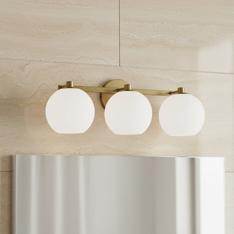 Enid 3 Light Vanity, Aged Brass - A three-globe vanity light fixture in aged brass with white glass shades is mounted on a travertine-tiled wall above a reflective mirror.