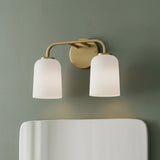 Elira 2 Light Vanity, Aged Brass - A brass vanity light with two white frosted glass shades is mounted on a green wall above a white mirror.