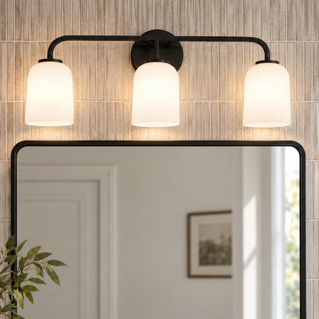 A transitional bathroom features the Elira 3 Light Vanity, Matte Black with frosted glass shades mounted above a rounded mirror on a beige fluted tile wall.